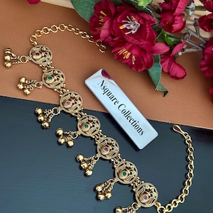 May include: Gold-toned headpiece with intricate circular designs, adorned with red and green gemstones and small dangling bells. The headpiece is connected by a delicate chain. The image also includes a white label with the text "Nsquare Collections".