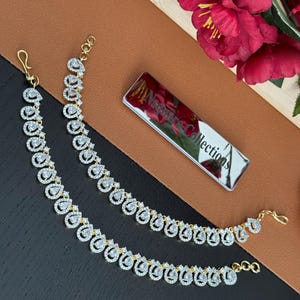 May include: Two gold-toned anklets with a teardrop design, each adorned with clear, sparkling stones. The anklets are displayed on a brown and black surface, with a mirrored tag that reads "Nsquare Collections" and red flowers.