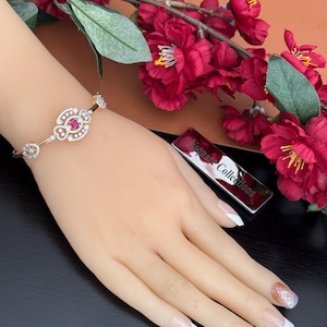 May include: A gold-toned bracelet with a central oval design featuring a red gemstone, surrounded by clear crystals. The bracelet is on a mannequin hand, with a silver rectangular object that reads "Vequare Collections".