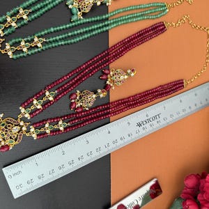 May include: An overhead shot showcases two necklaces and a pair of earrings. One necklace is green, the other is a deep red with gold accents. A ruler shows the necklace is approximately 30 inches long. The jewelry is on a black and orange background.