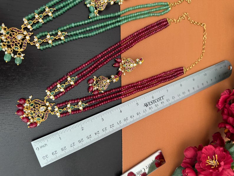 May include: An overhead shot showcases two necklaces and a pair of earrings. One necklace is green, the other is a deep red with gold accents. A ruler shows the necklace is approximately 30 inches long. The jewelry is on a black and orange background.