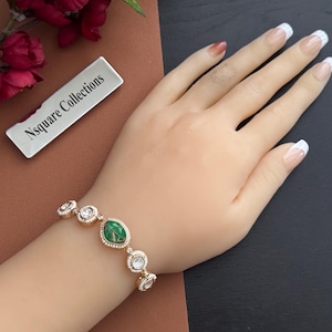 May include: A gold-toned bracelet featuring an oval green gemstone and several round, clear gemstones. The bracelet is adorned with small, clear crystals around each gemstone. The bracelet is on a person's wrist. The brand name "Nsquare Collections" is visible.