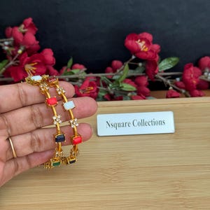 May include: Gold-toned bangles with colorful rectangular gemstones. The gemstones are red, white, blue, green, and black. The bangles are held in front of a wooden surface with red flowers in the background. The text "Nsquare Collections" is visible.