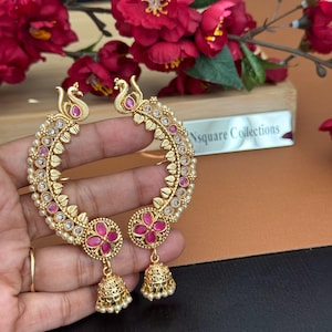 Peut inclure: Boucles d'oreilles dorées avec un motif de paon. Les boucles d'oreilles comportent des pierres précieuses roses, de petites perles blanches et des breloques en forme de clochettes pendantes. Les boucles d'oreilles sont tenues dans une main, avec des fleurs rouges en arrière-plan.