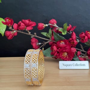 May include: Two gold bangles with detailed floral and geometric designs, embellished with small white stones. The bangles are presented on a wooden surface, with red flowers and a sign reading "Nsquare Collections" in the background.