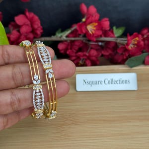 Puede incluir: Dos brazaletes dorados con intrincados detalles de piedras preciosas blancas. Los brazaletes se muestran sobre un fondo de flores rojas y una superficie de madera. El texto "Nsquare Collections" es visible.