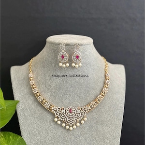 May include: A gold-coloured necklace and earring set on a grey mannequin. The necklace has a central floral design with a red gemstone and dangling pearls. The earrings mirror the design with red gemstones and pearls. The text "Nsquare Collections" is visible.