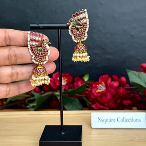 May include: Gold-coloured peacock earrings with red, white, and green details. The earrings showcase a detailed peacock design with a bell-shaped base decorated with small pearls. The earrings are displayed on a black stand against a dark backdrop.