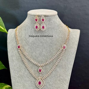 May include: A two-layer necklace and matching drop earrings set featuring clear stones in a leaf-like pattern with teardrop-shaped pink stones. The necklace has a central pendant and a smaller drop, with the text Nsquare Collections visible.