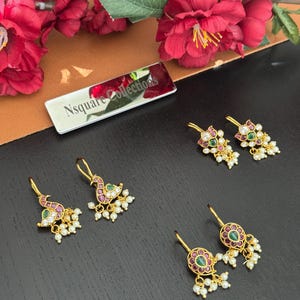 Op de afbeelding: Goudkleurige oorbellen met ingewikkelde ontwerpen, met rode, groene en witte edelstenen en kleine, bungelende parels. De oorbellen worden gepresenteerd op een donkere ondergrond, met een bloemstuk op de achtergrond. De oorbellen hebben een pauwen- en bloemenontwerp.