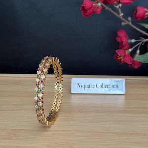 May include: A gold-toned bangle bracelet featuring a repeating pattern of clear, red, and green circular stones set in a metal frame. A small rectangular sign with the text Nsquare Collections is positioned to the right of the jewelry piece.