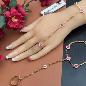 May include: Gold-toned hand jewelry set with ruby-colored stones. The set includes a ring, a bracelet, and a hand chain connecting the ring to the bracelet. The jewelry is displayed on a hand with a brown and black background.