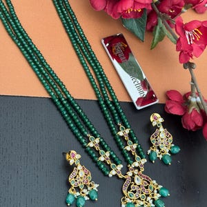 May include: Emerald green beaded necklace and matching earrings set. The necklace features multiple strands of small, round beads. The earrings and necklace pendants have gold-colored metal accents with red and clear stone details. The set is displayed with red flowers.