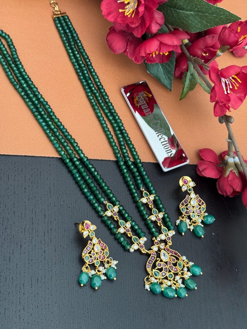 May include: Emerald green beaded necklace and matching earrings set. The necklace features multiple strands of small, round beads. The earrings and necklace pendants have gold-colored metal accents with red and clear stone details. The set is displayed with red flowers.