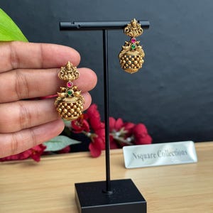 May include: Gold-toned earrings with intricate detailing. Each earring features a small figure at the top, a red and green gemstone, and a beaded, rounded base. The earrings are displayed on a black stand.