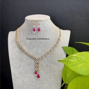 Może przedstawiać: Złoty naszyjnik i kolczyki z rubinowymi kamieniami w kształcie łezki i małymi, przezroczystymi kamieniami. Naszyjnik ma delikatny łańcuszek z wisiorkiem z trzech rubinowych łezek. Kolczyki odzwierciedlają wzór wisiorka. Widoczny jest tekst "Nsquare Collections".