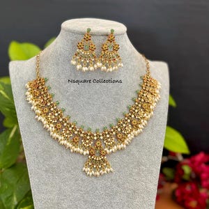 Può includere: Un set di collana e orecchini coordinati color oro. I gioielli presentano intricati disegni floreali, piccoli accenti verdi e rossi e perline bianche simili a perle. Il testo "Nsquare Collections" è visibile sul supporto espositivo.