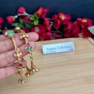 May include: Two gold-toned bangles featuring a twisted metal design, accented with small green and pink stones arranged in floral-like clusters. The bangles include dangling metal charms and a small white sign reading Nsquare Collections.