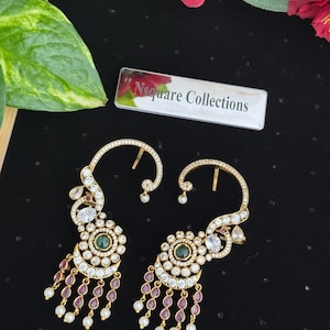 May include: Gold-coloured earrings with a floral design. Each earring has a central green stone surrounded by clear crystals, with dangling red and white accents. The earrings have a curved top and are set against a black background.