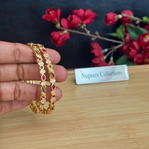 May include: Two gold-toned bangles featuring a repeating pattern of small clear stones, marquise-cut red stones, and round green stones. The metalwork includes intricate filigree details along the band, creating a textured and ornate appearance.