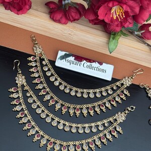 May include: Gold-toned, multi-layered ear chains with red and clear accents. Each layer features a teardrop design. The jewelry is displayed on a dark surface with a floral arrangement and a tag that reads "Nsquare Collections."
