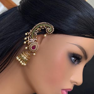 May include: An elaborate gold ear cuff with a green and white pattern, a central red gemstone, and a dangling bell-shaped charm. The cuff is decorated with small white pearls and clear crystals, offering a luxurious appearance.