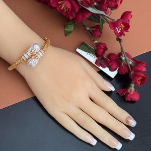 May include: Gold-toned bracelet with a pearl and rhinestone design. The bracelet features two large pearls and is adorned with sparkling rhinestones. The bracelet is displayed on a mannequin hand, with a background of red flowers and a brown and black surface.