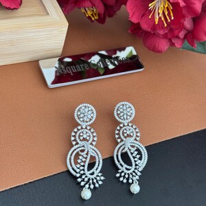 Pu&ograve; includere: Un paio di orecchini pendenti color argento con un design floreale. Ogni orecchino presenta tre elementi circolari adornati con cristalli trasparenti e una piccola perla bianca in basso. Gli orecchini sono esposti su una superficie marrone e nera.