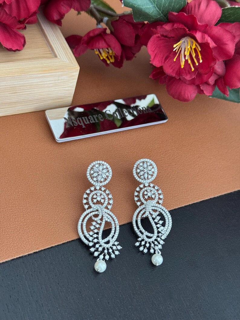 Pu&ograve; includere: Un paio di orecchini pendenti color argento con un design floreale. Ogni orecchino presenta tre elementi circolari adornati con cristalli trasparenti e una piccola perla bianca in basso. Gli orecchini sono esposti su una superficie marrone e nera.