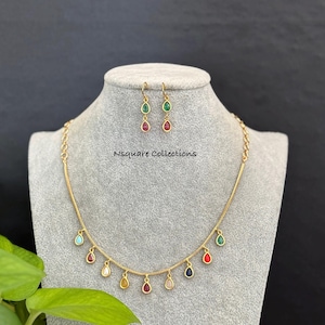 Puede incluir: Juego de collar y pendientes dorados. El collar presenta una delicada cadena con colgantes coloridos en forma de lágrima. Los pendientes tienen un diseño a juego. El texto "Nsquare Collections" es visible.