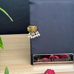 May include: Gold-toned earring with a rectangular top featuring intricate patterns and a cluster of small, white pearls hanging below. The earring is displayed on a black surface. The brand name "Nsquare Collections" is visible.