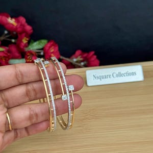 May include: Two gold-toned bangles with a delicate design. One bangle features a row of small, sparkling stones, while the other has a similar design with square accents. The bangles are displayed against a backdrop of red flowers and a wooden surface.