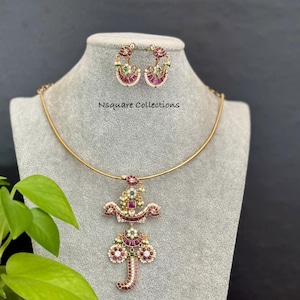 May include: A gold-coloured necklace and earring set. The necklace has a pendant with red, green, and white accents. The earrings are half-hoop style, with similar embellishments. The text "Nsquare Collections" is visible.