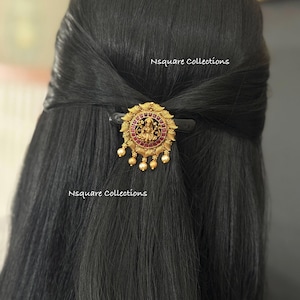 May include: A gold-colored hair clip with a circular pendant featuring a central figure, surrounded by red stones and gold detailing. The clip is holding back dark hair. The text "Nsquare Collections" is visible.