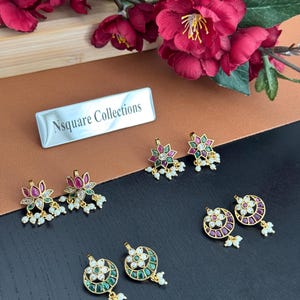 Op de afbeelding: Een assortiment goudkleurige oorbellen met kleurrijke edelstenen en kleine witte parels. De oorbellen hebben verschillende bloemen- en geometrische ontwerpen. De afbeelding bevat een bord met de tekst "Nsquare Collections."