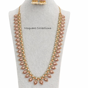 May include: A gold-coloured necklace and matching earrings set. The necklace has a repeating pattern of red and white floral-shaped pendants with green accents. The earrings have a similar design with pearl drops. The text "Nsquare Collections" is visible.