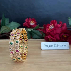 May include: Gold-toned bangle bracelet with alternating ruby and emerald-colored stones. The bracelet has a floral design and is displayed on a wooden surface. The text "Nsquare Collections" is visible in the background.