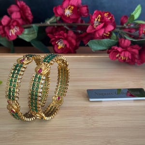 以下が含まれることがあります： エメラルドグリーンとルビーレッドのアクセントが施された2つのゴールドバングル。バングルには小さな白い宝石と複雑なディテールが施されています。背景には赤い花と木製の表面があります。