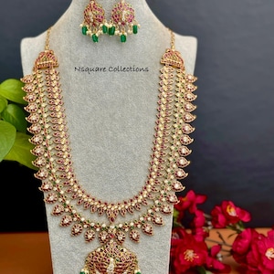 May include: A gold-toned necklace and earring set with red and green gemstones. The necklace has a long, ornate design with a pendant. The earrings are matching, with a similar design. The set is displayed on a gray mannequin.