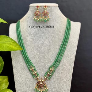 May include: A green beaded necklace and matching earrings set. The necklace features a pendant with red and green accents. The earrings have a similar design, with green beads and gold-colored metal. The text "Nsquare Collections" is visible.