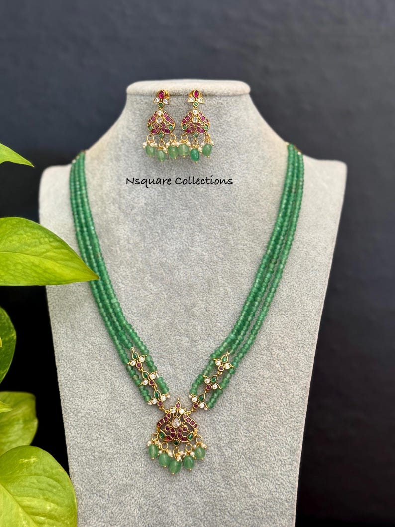 May include: A green beaded necklace and matching earrings set. The necklace features a pendant with red and green accents. The earrings have a similar design, with green beads and gold-colored metal. The text "Nsquare Collections" is visible.