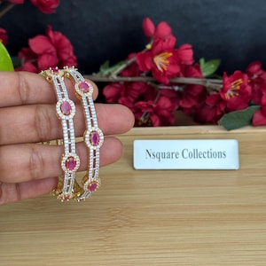 Puede incluir: Dos brazaletes dorados con piedras de color rubí ovaladas y piedras de acento redondas y transparentes. Los brazaletes se muestran sobre un fondo oscuro con flores rojas y una superficie de madera. El texto "Nsquare Collections" es visible.