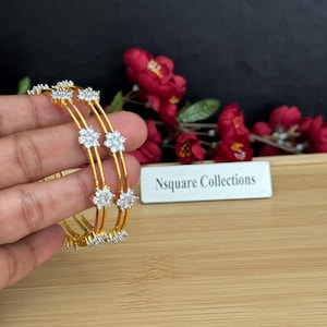 May include: Gold-toned bangles with floral accents. Each bangle features a series of small, sparkling flower-shaped embellishments. The bangles are held against a backdrop of red flowers and a wooden surface. The text "Nsquare Collections" is visible.