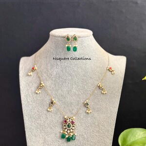 May include: Gold-toned necklace and earring set with emerald green and ruby red accents. The necklace features a pendant with a central green stone and dangling green teardrop-shaped stones. The earrings have a similar design. The text "Nsquare Collections" is visible.