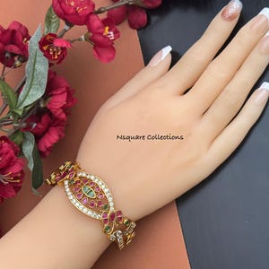 May include: A gold-toned bracelet with an oval centerpiece, adorned with red, green, and clear gemstones. The bracelet is on a wrist, with the text "Nsquare Collections" visible. Red flowers are in the background.