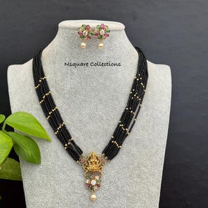 May include: A black beaded necklace and matching earrings set. The necklace features multiple strands of black beads with gold accents and a gold pendant with a deity figure. The earrings are flower-shaped with colorful gemstones and pearl drops. The text "Nsquare Collections" is visible.