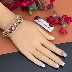 Puede incluir: Una pulsera dorada con un diseño floral, con piedras preciosas rojas, verdes y blancas y perlas. La pulsera se muestra en una mano de maniquí. El fondo presenta flores rojas y una etiqueta plateada con el texto "square Collections".