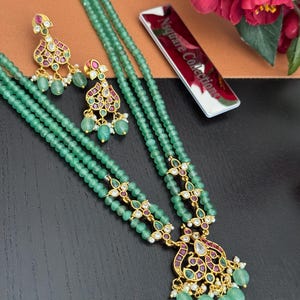 May include: A three-strand green beaded necklace and matching earrings set. The necklace features gold-toned filigree accents with red and purple gemstones. The earrings have a similar design, with green beads and small white pearls.