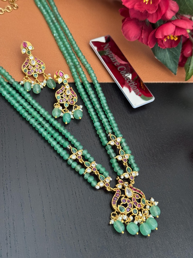 May include: A three-strand green beaded necklace and matching earrings set. The necklace features gold-toned filigree accents with red and purple gemstones. The earrings have a similar design, with green beads and small white pearls.