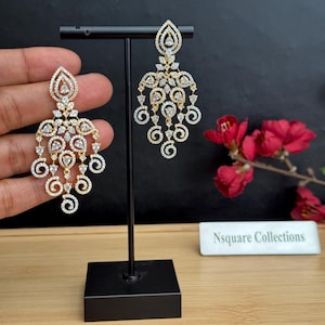 May include: A pair of gold-tone chandelier earrings featuring clear, pear-shaped and round-cut stones arranged in intricate swirling patterns. The earrings are displayed on a black stand, with a sign reading Nsquare Collections nearby.